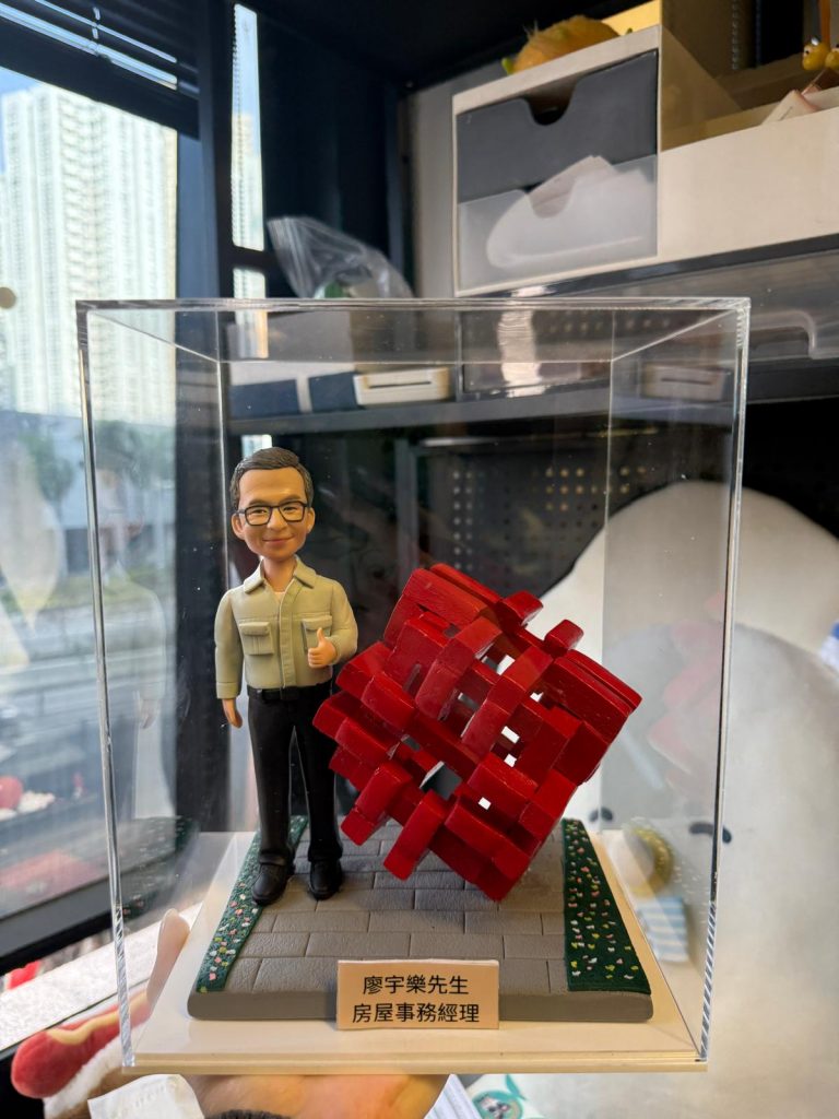 透明展示盒中的客製公仔站在灰色地台上，身穿淺色襯衫與黑色長褲，右手比讚；旁邊有一個紅色編織感的幾何立方體雕塑；地台前方有金色小名牌刻有中文職稱與姓名，四周點綴綠色草地與小花紋路；背景為室內辦公與窗景。