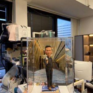 透明展示盒中的客製公仔，身穿深色西裝，右手握著麥克風站立；腳下為藍金圖案小基座，背板是一條金色與石牆相間的室內走廊影像；前方金色名牌上印有中文祝福字樣；手持展示盒的人位於辦公室環境中。