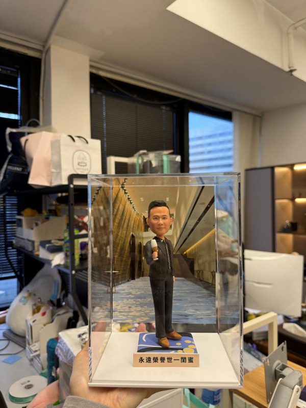 透明展示盒中的客製公仔，身穿深色西裝，右手握著麥克風站立；腳下為藍金圖案小基座，背板是一條金色與石牆相間的室內走廊影像；前方金色名牌上印有中文祝福字樣；手持展示盒的人位於辦公室環境中。