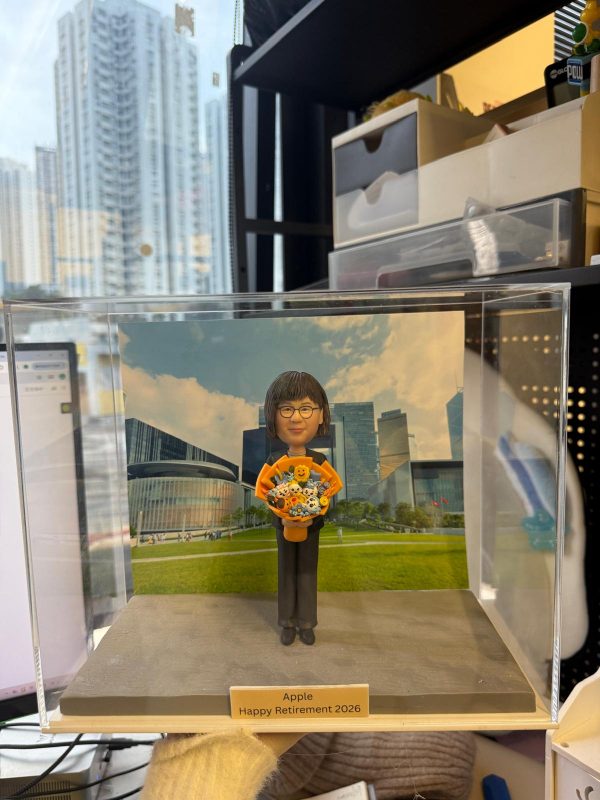 透明展示盒中的客製公仔，身穿黑色長褲與橘色元素的上衣，雙手捧著卡通造型花束；背板是草地與現代建築群的風景圖；前方金色小名牌寫有「Apple Happy Retirement 2026」；擺放在室內桌面前，背景可見辦公室收納物與窗外高樓。