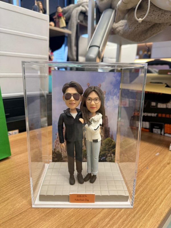 透明展示盒內的雙人客製公仔並肩站立，左側公仔穿深色外套與太陽眼鏡，右側公仔穿白色外套與淺色長褲；腳下為灰色方格地台，背板為歐式教堂風景照；前方橘色小名牌印有 2026.02.14 Valentine’s Day 字樣；擺放在木質桌面，背景為室內陳列與收納物品。