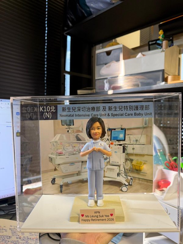 透明展示盒中的醫護主題客製公仔，穿著淺藍色護理服與白鞋，雙手在胸前比出愛心手勢；站在淺色小台座上，前方名牌寫有「APN Ms Leung Suk Yee Happy Retirement 2026」並配有紅色愛心；背板是新生兒加護病房設備與保溫箱的場景，展示盒上緣貼有「新生兒深切治療部及 新生兒特別護理部／Neonatal Intensive Care Unit & Special Care Baby Unit」字樣；背景為室內辦公環境。