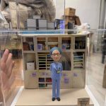 透明展示盒中的醫護主題客製公仔，穿淡藍色制服與頭套、配識別證，雙手在身後站立；背後是分類櫃與文件層架的微縮場景，貼有多張小標籤；前方右側立有金色小銘牌，整體置於木桌與室內背景前。