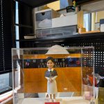 透明展示盒中的客製公仔，穿白色連身裙與深色披肩，雙手交握站在方形底座；背板為木質色並貼有「綜合腎科中心 INTEGRATED RENAL CENTRE」字樣與心形圖示，底座前有紅色小名牌寫著祝賀退休訊息；周圍可見辦公室置物架與雜物。