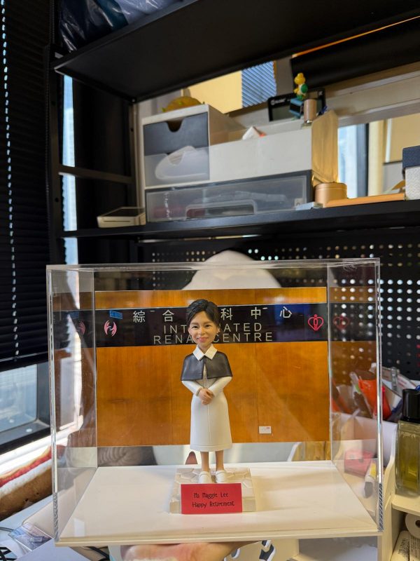 透明展示盒中的客製公仔，穿白色連身裙與深色披肩，雙手交握站在方形底座；背板為木質色並貼有「綜合腎科中心 INTEGRATED RENAL CENTRE」字樣與心形圖示，底座前有紅色小名牌寫著祝賀退休訊息；周圍可見辦公室置物架與雜物。