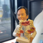 穿白色實驗袍的醫療主題客製公仔，雙手托著帶底座的人體腸道模型，模型上標示多處紅色病變；背景為室內辦公與窗邊場景。