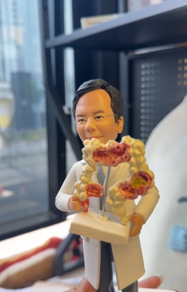 穿白色實驗袍的醫療主題客製公仔，雙手托著帶底座的人體腸道模型，模型上標示多處紅色病變；背景為室內辦公與窗邊場景。