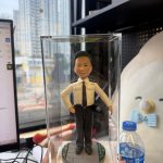 手持透明展示盒的近景照：盒內是一位身穿淺色制服襯衫、黑領帶與黑長褲的警務主題公仔，右手叉腰、左手持警帽；腳下為圓形灰色地磚基座，邊緣點綴綠地與小花紋；背景為辦公室桌面與窗外高樓，右側可見一瓶礦泉水。