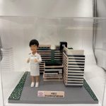 透明展示盒內的醫療主題客製公仔，身穿白色制服與白鞋、雙手比讚站在灰色地台上；旁邊有縮小版建築與花園模型，前方小牌寫有中文姓名與「深切治療部門運作經理」等字樣並帶有紅色徽標；地台兩側有綠色草地與小花紋路；背景為白色布幕。