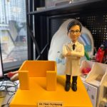 手持黃色台座的醫療主題客製公仔：公仔穿白袍、黑褲與黑鞋，雙手交叉站立在右側；台座左側有一個開放式方形結構；前方白色名牌以中英文字樣標示「Dr. Tien-Yu Huang／黃天祐 醫師」。背景為室內辦公與收納架、窗景。