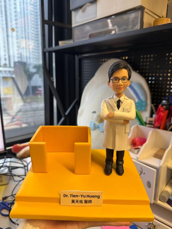 手持黃色台座的醫療主題客製公仔：公仔穿白袍、黑褲與黑鞋，雙手交叉站立在右側；台座左側有一個開放式方形結構；前方白色名牌以中英文字樣標示「Dr. Tien-Yu Huang／黃天祐 醫師」。背景為室內辦公與收納架、窗景。