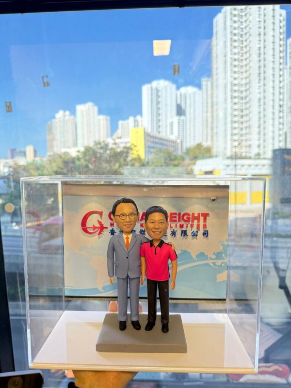 透明展示盒中的雙人客製公仔並肩站立，左側穿淺灰西裝配橘色領帶，右側穿桃紅與黑色拼接的休閒上衣與黑長褲；腳下為灰色長方形基座；背板印有公司英文縮寫與中文公司名稱的圖樣；背景透過玻璃可見高樓林立的城市景色與藍天。