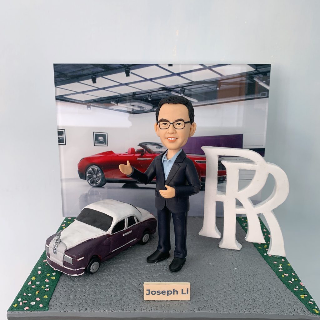 透明展示盒內的客製男士公仔,穿深色西裝站在迷你勞斯萊斯車旁,背板為豪華車展示廳,旁立大型「RR」字母,銘牌寫「Joe Li」
