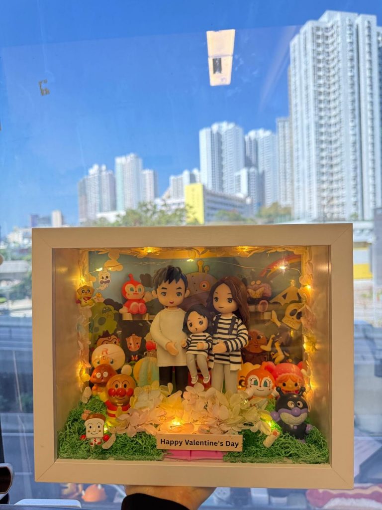 白色框的立體燈箱相框內放置一家三口黏土公仔，前景鋪有綠色草粉與粉白色花材，邊緣繞暖黃小燈串，底部木牌寫著「Happy Valentine’s Day」。背景板為多位可愛卡通角色與彩虹圖案，可見麵包超人（Anpanman）、細菌人（Baikinman）等。相框置於木桌或在窗前手持拍攝，室內辦公室陳設、窗外高樓作為背景。