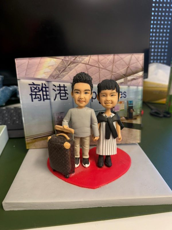 CUTEFIGUREHK 客製化情侶人像公仔，男女主角站在紅色心形底座上，男主角手扶精緻行李箱，配搭香港國際機場「離港 Departures」背景板，是專為親友送行訂製的移民禮物與送別禮物。
