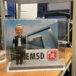 CUTEFIGUREHK 客製化西裝男士人像公仔，公仔擺出派心手勢，配搭 EMSD 機電工程署標誌名牌及總部大樓背景的透明展示盒，是專為工程界前輩訂製的退休禮物與工程禮物。