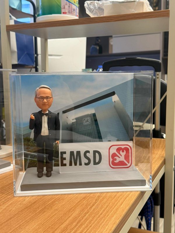 CUTEFIGUREHK 客製化西裝男士人像公仔，公仔擺出派心手勢，配搭 EMSD 機電工程署標誌名牌及總部大樓背景的透明展示盒，是專為工程界前輩訂製的退休禮物與工程禮物。