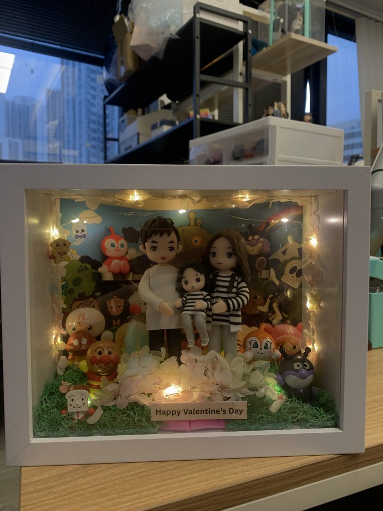 白色框的立體燈箱相框內放置一家三口黏土公仔，前景鋪有綠色草粉與粉白色花材，邊緣繞暖黃小燈串，底部木牌寫著「Happy Valentine’s Day」。背景板為多位可愛卡通角色與彩虹圖案，可見麵包超人（Anpanman）、細菌人（Baikinman）等。相框置於木桌或在窗前手持拍攝，室內辦公室陳設、窗外高樓作為背景。