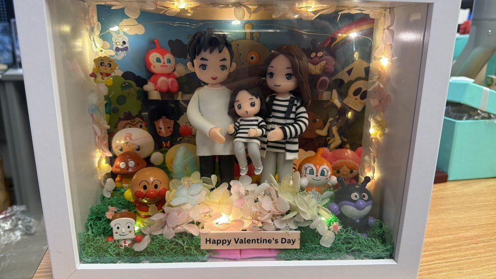 白色框的立體燈箱相框內放置一家三口黏土公仔，前景鋪有綠色草粉與粉白色花材，邊緣繞暖黃小燈串，底部木牌寫著「Happy Valentine’s Day」。背景板為多位可愛卡通角色與彩虹圖案，可見麵包超人（Anpanman）、細菌人（Baikinman）等。相框置於木桌或在窗前手持拍攝，室內辦公室陳設、窗外高樓作為背景。