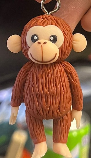 近拍棕色猴子造型鑰匙圈公仔：頭頂連著金屬吊環，圓耳、大眼與米色嘴鼻區，身體有立體毛流紋理，手腳為淺色，背景散景模糊。