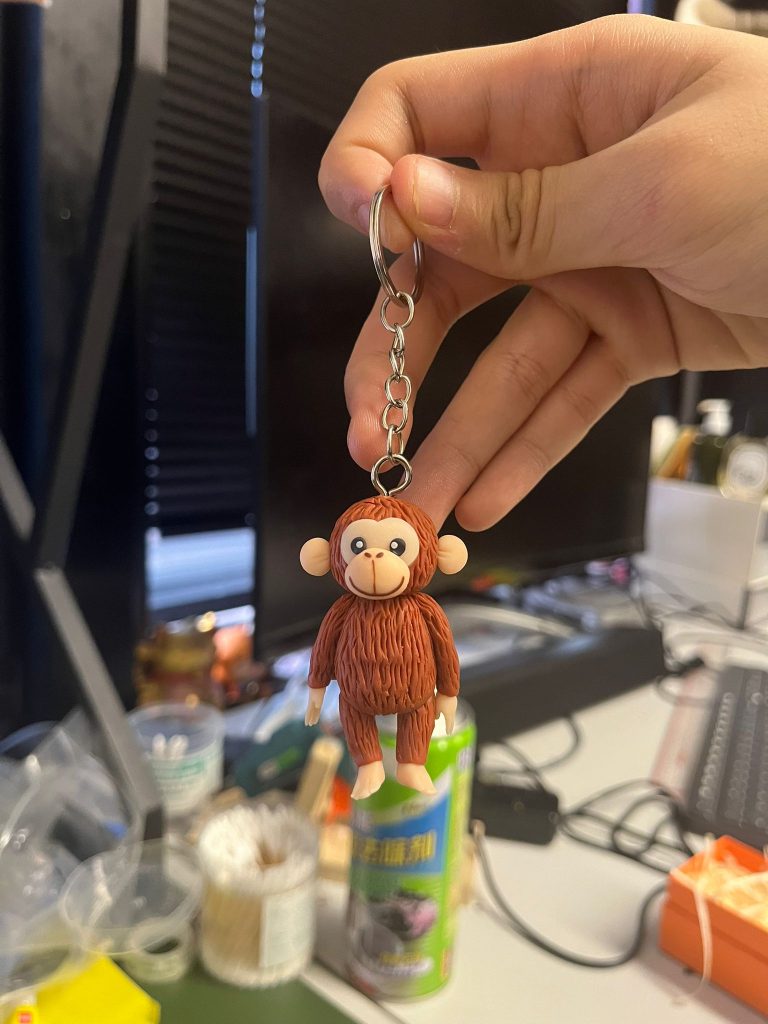 近拍棕色猴子造型鑰匙圈公仔：頭頂連著金屬吊環，圓耳、大眼與米色嘴鼻區，身體有立體毛流紋理，手腳為淺色，背景散景模糊。