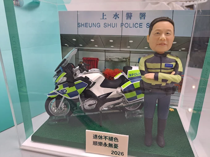 透明展示盒內的警務主題公仔場景:穿著螢光黃與深色相間制服的公仔雙手交叉站立,旁邊是一台貼有藍綠方格反光貼的白色警用大型機車;背板印有「SHEUNG SHUI POLICE S…」及中文所名;綠色草皮底座前方有金屬色名牌,刻有祝福與「2026」字樣。