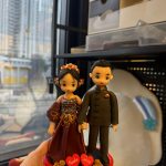 CUTEFIGUREHK 客製化結婚情侶人像公仔。女方身穿深紅色鑲金邊華麗晚裝並配戴精緻花卉頭飾，男方身穿黑色中式立領禮服。兩人甜蜜牽手站立於喜慶的紅色圓形底座上，底座飾有立體金色玫瑰花，並帶有寫著新人名字「Nadia」和「Jefrey」的紅心，以及「Happy Wedding」字樣的紅色牌匾。這是一份極具紀念價值的專屬結婚禮物。