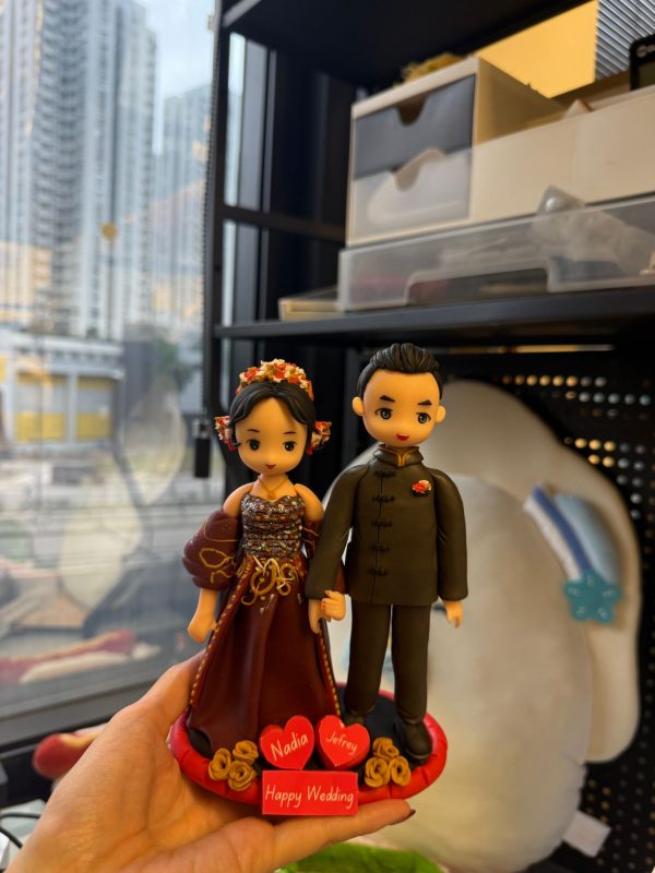 CUTEFIGUREHK 客製化結婚情侶人像公仔。女方身穿深紅色鑲金邊華麗晚裝並配戴精緻花卉頭飾，男方身穿黑色中式立領禮服。兩人甜蜜牽手站立於喜慶的紅色圓形底座上，底座飾有立體金色玫瑰花，並帶有寫著新人名字「Nadia」和「Jefrey」的紅心，以及「Happy Wedding」字樣的紅色牌匾。這是一份極具紀念價值的專屬結婚禮物。
