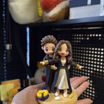 CUTEFIGUREHK 客製化情侶禮物公仔。一隻手正托著一個精緻的立體雙人公仔，背景是擺放了毛公仔的層架。公仔是一對情侶，擺出電影《鐵達尼號》(Titanic) 的經典船頭飛翔姿勢。男主角在後方環抱女主角的腰部，女主角雙手向兩側平舉。兩人都穿著深色長外套搭配金色袖口，女主角內搭白色連身裙。公仔站在仿沙灘質感的底座上，底座前方有一個藍黃相間的救生圈和一枚白色小貝殼。