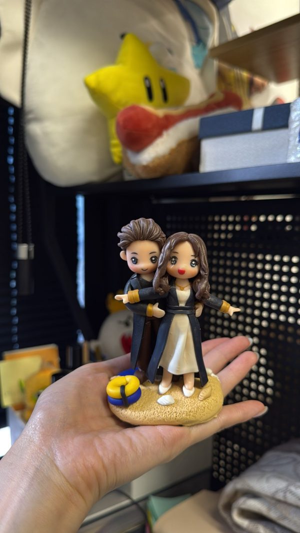 CUTEFIGUREHK 客製化情侶禮物公仔。一隻手正托著一個精緻的立體雙人公仔，背景是擺放了毛公仔的層架。公仔是一對情侶，擺出電影《鐵達尼號》(Titanic) 的經典船頭飛翔姿勢。男主角在後方環抱女主角的腰部，女主角雙手向兩側平舉。兩人都穿著深色長外套搭配金色袖口，女主角內搭白色連身裙。公仔站在仿沙灘質感的底座上，底座前方有一個藍黃相間的救生圈和一枚白色小貝殼。