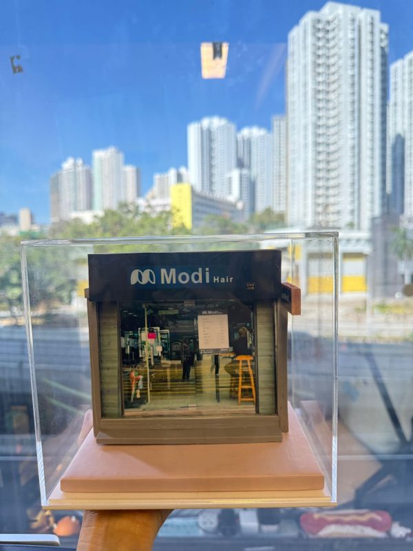 CUTEFIGUREHK 客製化店舖微縮模型。一個精緻的微型髮型屋店面模型，頂部有黑底藍字及白字的招牌寫著「Modi Hair」和「安寧路49號」。店面採用透明玻璃門設計，內部貼有價目表海報，隱約可見店內的裝潢和一張黃色高腳椅。模型放置在一個淺粉紅色的方形底座上，並罩著透明的壓克力展示盒。背景是隔著玻璃窗看到的城市高樓大廈和藍天。