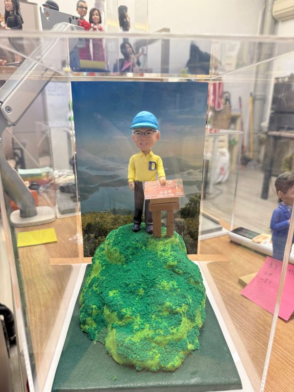 CUTEFIGUREHK 客製化行山主題紀念公仔。一位戴著淺藍色鴨舌帽和眼鏡的男士，身穿胸前印有藍色英文字母「D」的黃色Polo恤和深色長褲，面帶笑容地站在一個立體的綠色草地山頂模型上。他身旁立著一塊寫有「仙姑峰」（Hsien Ku Fung）及海拔高度的仿真木牌。公仔背後是一幅壯麗的香港山水風景（八仙嶺及船灣淡水湖）背景圖。整個模型被精心放置在一個透明的亞加力膠展示盒中。