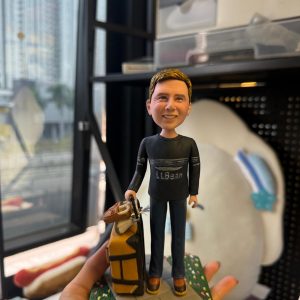 CUTEFIGUREHK 客製化男士人像公仔，生動還原了身穿深色 L.L.Bean 上衣及牛仔褲的男士，手拖著放有旅行袋的黃色行李箱。公仔站立於帶有草地小徑的底座上，前方設有寫著「Heartfelt thanks for being our great leader」的專屬感謝牌。這是一份極具心思、專為感謝上司或領導而設的散水、升職或榮休紀念禮物。
