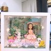 CUTEFIGUREHK 客製化立體相框公仔禮物。一個白色的發光畫框，內部佈置成夢幻的童話花園。中央是一個穿著粉紅色花瓣晚裝的長髮女孩立體公仔，頭戴花環，手持粉紅玫瑰。背景是浪漫的黃昏花園圖案，公仔腳下鋪滿了紫色、白色和粉紅色的乾花（繡球花）以及綠色苔蘚，旁邊點綴著微縮的白色小噴泉、可愛的小棕熊和熊貓模型。相框內置有溫馨的 LED 燈串，營造出唯美的氣氛。