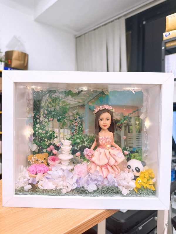 CUTEFIGUREHK 客製化立體相框公仔禮物。一個白色的發光畫框，內部佈置成夢幻的童話花園。中央是一個穿著粉紅色花瓣晚裝的長髮女孩立體公仔，頭戴花環，手持粉紅玫瑰。背景是浪漫的黃昏花園圖案，公仔腳下鋪滿了紫色、白色和粉紅色的乾花（繡球花）以及綠色苔蘚，旁邊點綴著微縮的白色小噴泉、可愛的小棕熊和熊貓模型。相框內置有溫馨的 LED 燈串，營造出唯美的氣氛。