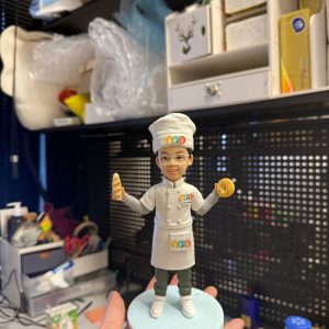 CUTEFIGUREHK 客製化兒童廚師公仔。一個戴著金屬圓框眼鏡的小男孩，身穿專業的白色廚師服、高帽及圍裙，制服上印有彩色的廚藝學校標誌。他笑容燦爛，右手拿著法式長棍麵包，左手拿著貝果（Bagel），下身穿著深綠色長褲及白鞋，站在淺藍色的圓形底座上。這是一份送給熱愛烹飪的小朋友的完美禮物。