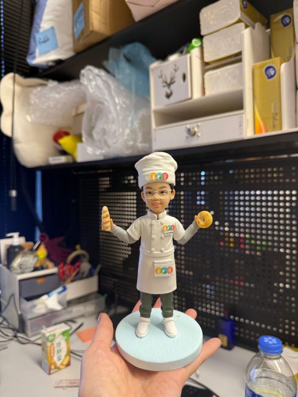 CUTEFIGUREHK 客製化兒童廚師公仔。一個戴著金屬圓框眼鏡的小男孩，身穿專業的白色廚師服、高帽及圍裙，制服上印有彩色的廚藝學校標誌。他笑容燦爛，右手拿著法式長棍麵包，左手拿著貝果（Bagel），下身穿著深綠色長褲及白鞋，站在淺藍色的圓形底座上。這是一份送給熱愛烹飪的小朋友的完美禮物。
