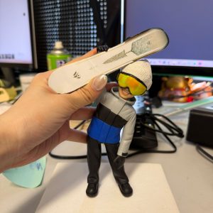 CUTEFIGUREHK 客製化滑雪造型禮物公仔。一隻手正拿著一個精緻的立體真人公仔，背景是辦公桌和電腦螢幕。公仔主角是一位笑容滿面的男士，頭戴白色冷帽和搶眼的黃色反光滑雪鏡，身穿灰、黑、藍拼色的滑雪外套及黑色雪褲。他單手高舉著一塊印有綠色樹木圖案的白色單板滑雪板 (Snowboard)。公仔站在白色的方形底座上，底座前方有一塊藍色名牌，上面用黑字寫著生日祝福「Andrew Happy Birthday」。