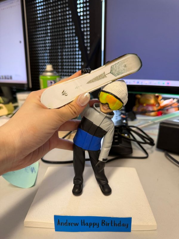 CUTEFIGUREHK 客製化滑雪造型禮物公仔。一隻手正拿著一個精緻的立體真人公仔，背景是辦公桌和電腦螢幕。公仔主角是一位笑容滿面的男士，頭戴白色冷帽和搶眼的黃色反光滑雪鏡，身穿灰、黑、藍拼色的滑雪外套及黑色雪褲。他單手高舉著一塊印有綠色樹木圖案的白色單板滑雪板 (Snowboard)。公仔站在白色的方形底座上，底座前方有一塊藍色名牌，上面用黑字寫著生日祝福「Andrew Happy Birthday」。