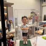 一尊裝在透明壓克力展示盒內的客製化男性足球員立體公仔。公仔雙手交叉於胸前，身穿印有「GIN BOUTIQUE」字樣與隊徽的白色足球球衣、黑色短褲（褲管印有數字14）以及紅白相間的球鞋。公仔站在綠色仿草皮質感的底座上，展現出運動員的自信風采。背景為擺放著其他公仔作品（如婚紗公仔）的工作室環境。