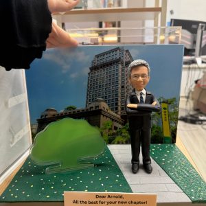 CUTEFIGUREHK 客製化企業紀念公仔。公仔為一位銀髮、戴眼鏡、身穿深色西裝打領帶的男士，雙手交叉抱胸，展現專業自信。場景配有國泰金控大樓背景圖及標誌性的綠色大樹道具。底座名牌刻有「Dear Arnold, All the best for your new chapter! Cathay United Bank Hong Kong Branch」，是一份專為銀行高管訂製的榮休或散水禮物。