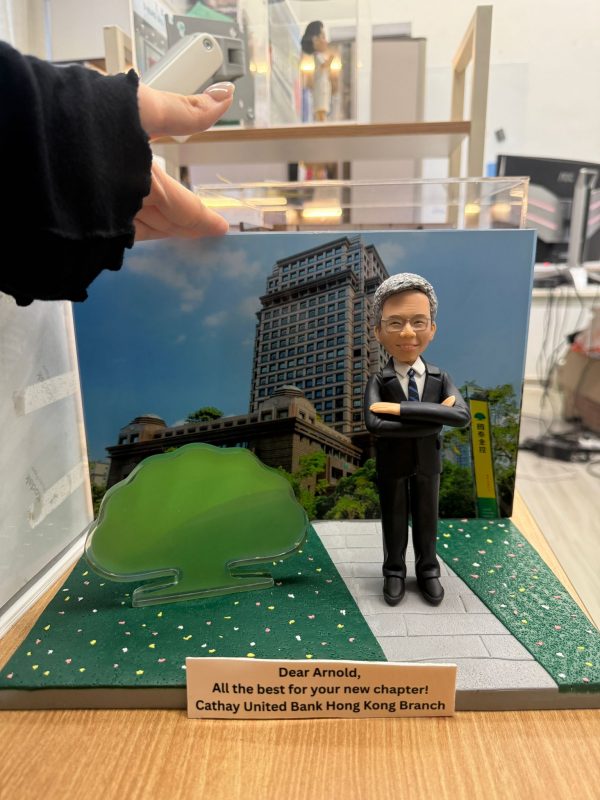 CUTEFIGUREHK 客製化企業紀念公仔。公仔為一位銀髮、戴眼鏡、身穿深色西裝打領帶的男士，雙手交叉抱胸，展現專業自信。場景配有國泰金控大樓背景圖及標誌性的綠色大樹道具。底座名牌刻有「Dear Arnold, All the best for your new chapter! Cathay United Bank Hong Kong Branch」，是一份專為銀行高管訂製的榮休或散水禮物。