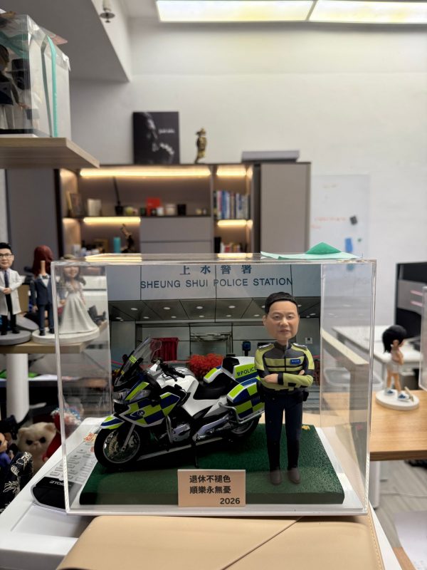 CUTEFIGUREHK 客製化香港交通警公仔。公仔身穿螢光黃綠色交通警制服，雙手交叉抱胸。旁邊配有一架精緻的警察電單車模型。場景背景為「上水警署」(Sheung Shui Police Station)。底座名牌刻有「退休不褪色 順樂永無憂 2026」，是一份專為警察訂製的榮休紀念禮物，裝在透明亞加力展示盒中。