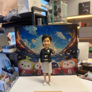 CUTEFIGUREHK 客製化女性主管人像公仔，生動還原了身穿黑色西裝外套、白色百褶裙及配戴員工證的女士造型，並擺出充滿自信的讚好（Thumbs up）手勢。公仔背後配有色彩繽紛的運動場背景，以及四隻印有「2026」字樣的港鐵吉祥物「鐵仔」、「機鐵仔」、「高鐵隊長」、「綠鐵B」公仔圖案。底座設有專屬牌匾，印有「Denise Kee, General Manager - Learning & HR Transformation, Best wishes on your new chapter in life!」的祝福語。這是一份極具心思的企業散水、榮休或升職紀念禮物。