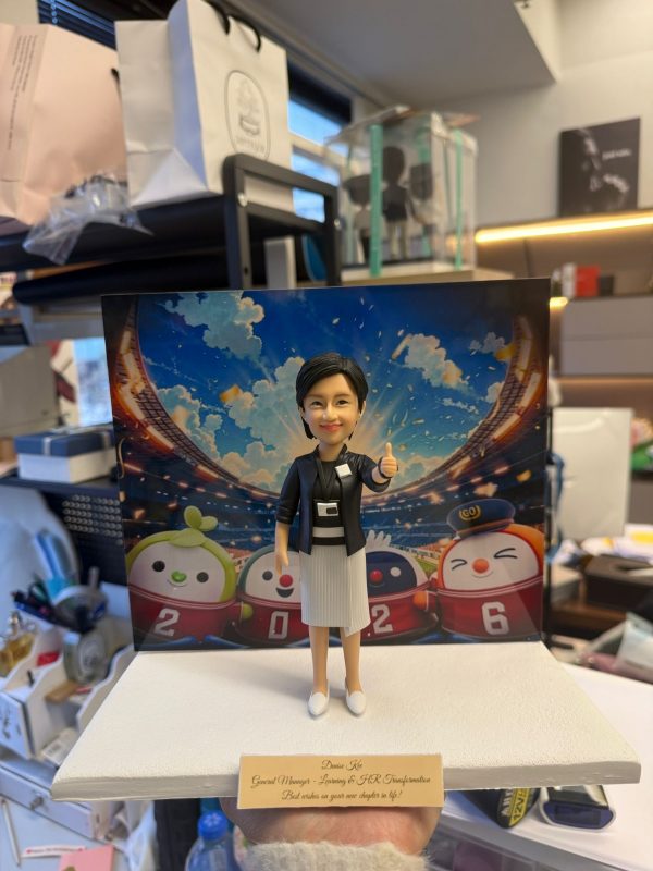 CUTEFIGUREHK 客製化女性主管人像公仔，生動還原了身穿黑色西裝外套、白色百褶裙及配戴員工證的女士造型，並擺出充滿自信的讚好（Thumbs up）手勢。公仔背後配有色彩繽紛的運動場背景，以及四隻印有「2026」字樣的港鐵吉祥物「鐵仔」、「機鐵仔」、「高鐵隊長」、「綠鐵B」公仔圖案。底座設有專屬牌匾，印有「Denise Kee, General Manager - Learning & HR Transformation, Best wishes on your new chapter in life!」的祝福語。這是一份極具心思的企業散水、榮休或升職紀念禮物。