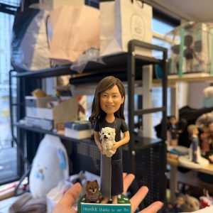 CUTEFIGUREHK 客製化真人與寵物紀念公仔。公仔造型為一位長髮微笑的女士，身穿黑灰拼接連身裙，懷中抱著一隻白色毛茸茸的小狗，腳邊還有一隻棕色小狗。綠色草地造型底座前方的名牌刻有「Heartfelt thanks to our Mei」，是一份充滿心意與感謝的專屬客製化禮物。
