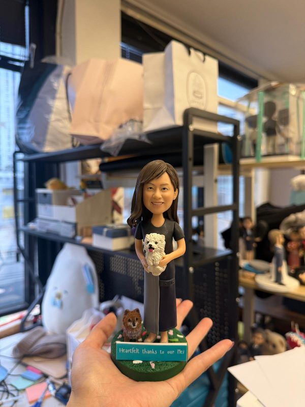 CUTEFIGUREHK 客製化真人與寵物紀念公仔。公仔造型為一位長髮微笑的女士，身穿黑灰拼接連身裙，懷中抱著一隻白色毛茸茸的小狗，腳邊還有一隻棕色小狗。綠色草地造型底座前方的名牌刻有「Heartfelt thanks to our Mei」，是一份充滿心意與感謝的專屬客製化禮物。