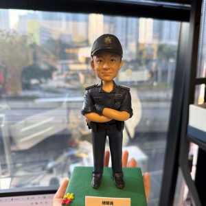 CUTEFIGUREHK 客製化香港海關人員人像公仔。公仔生動還原了人物的面貌，並穿著整齊的深藍色海關制服與海關帽，雙手交叉胸前，展現專業自信的姿態。公仔站立於點綴了小花的綠色底座上，前方設有印著「總關員 黃文傑 榮休之喜」的專屬紀念牌。這是一份專為紀律部隊人員度身訂造的退休致敬禮物。