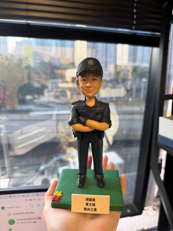 CUTEFIGUREHK 客製化香港海關人員人像公仔。公仔生動還原了人物的面貌，並穿著整齊的深藍色海關制服與海關帽，雙手交叉胸前，展現專業自信的姿態。公仔站立於點綴了小花的綠色底座上，前方設有印著「總關員 黃文傑 榮休之喜」的專屬紀念牌。這是一份專為紀律部隊人員度身訂造的退休致敬禮物。