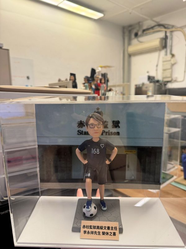 CUTEFIGUREHK 客製化榮休紀念公仔。一位戴著眼鏡的男士，身穿黑色運動套裝（上衣印有心形圖案及數字「168」，短褲亦印有「168」），雙手叉腰，右腳踩著一個足球，站立在仿石磚紋理的灰色底座上。底座前方設有一塊專屬名牌，印有「赤柱監獄高級文書主任 李永祥先生 榮休之喜」。公仔背後配有一幅印有香港懲教署徽章及「赤柱監獄 Stanley Prison」字樣的建築物背景圖，整體安放在一個透明的亞加力膠展示盒內。